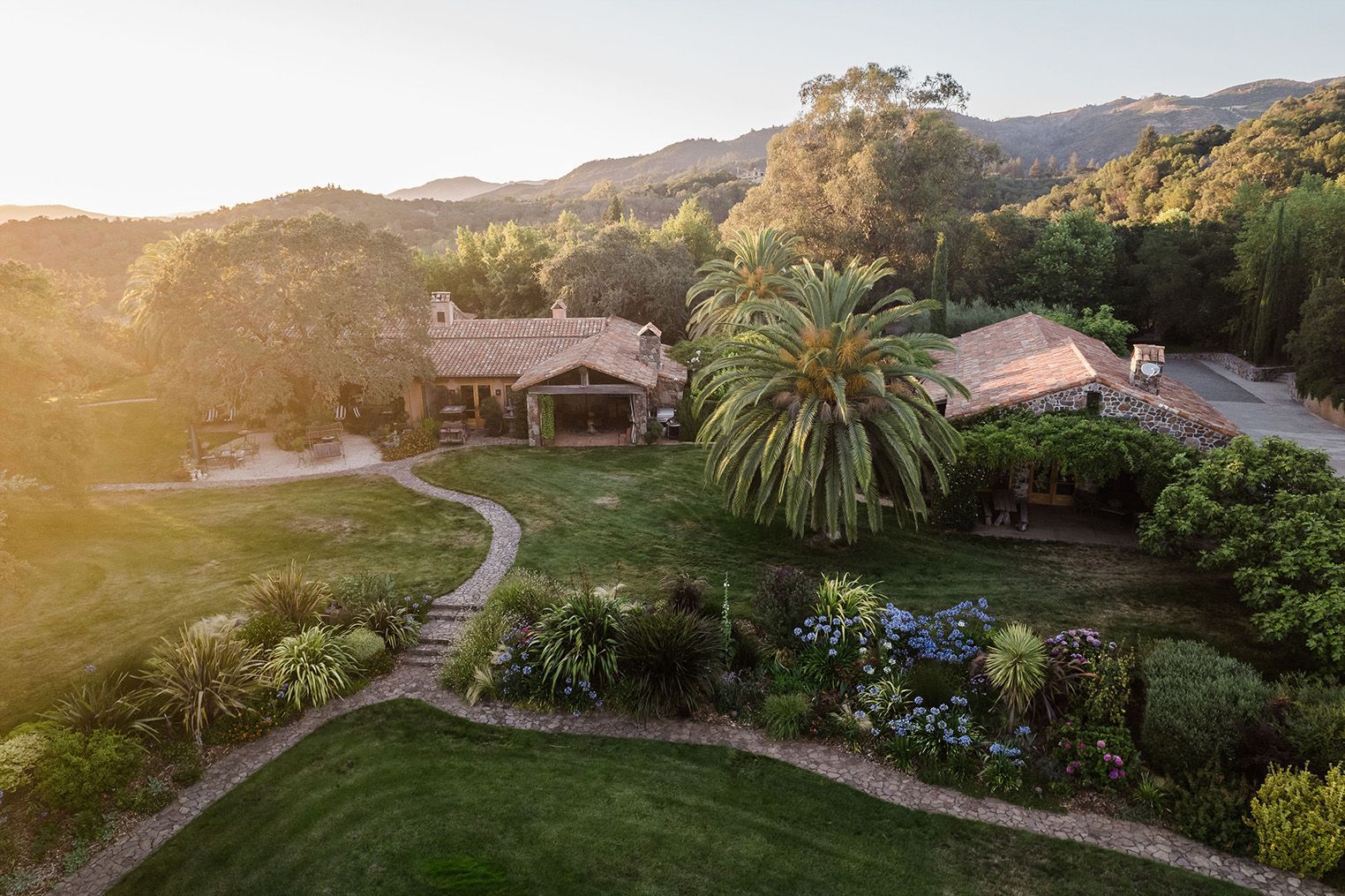 Sunlit hillside garden with winding path, palm tree, and rustic houses amid lush greenery