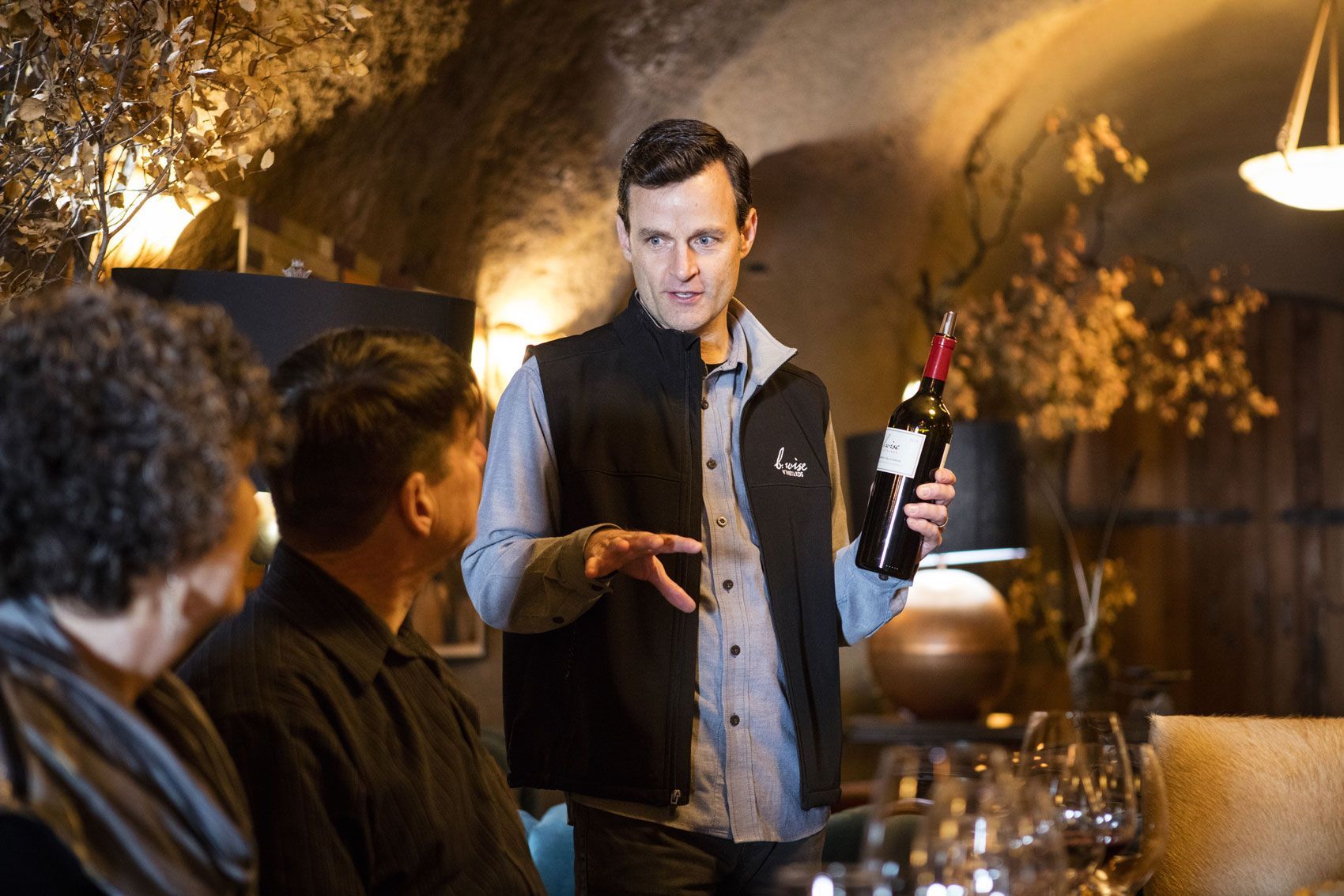 Man speaking and gesturing in a warmly lit bar, holding a bottle near seated guests.