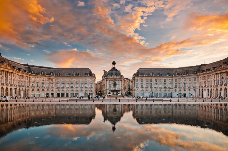 Grand historic palace with curved wings at sunset, reflected in still water under a colorful sky.