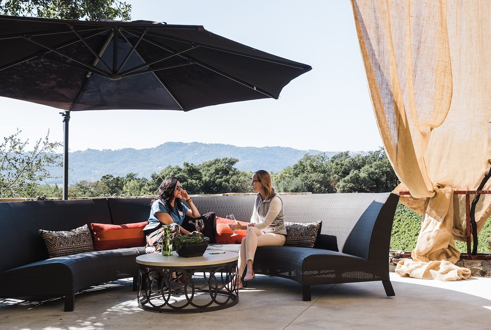Two people seated on a wicker patio sofa under a large umbrella, with mountains in the background.