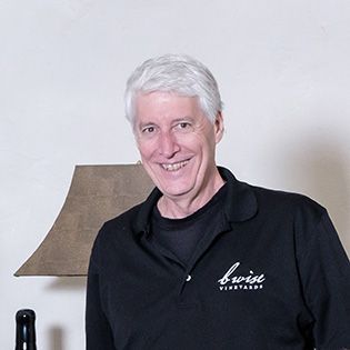 Smiling person in a black shirt standing indoors beside a table lamp