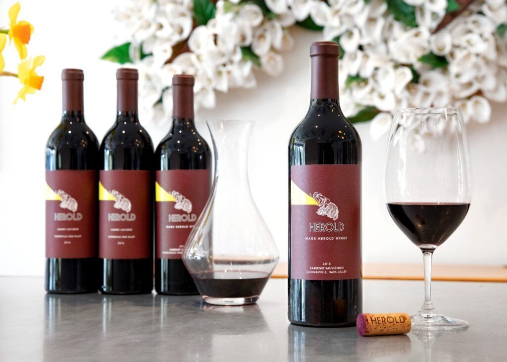 Four red wine bottles with glasses on a table, against a white floral backdrop