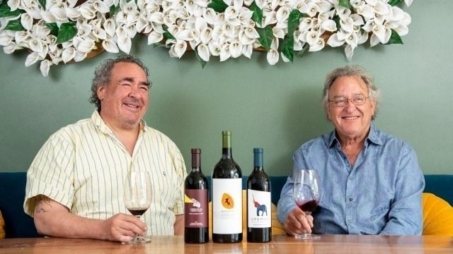 Two men smiling at a table with wine bottles and glasses in front of a floral backdrop