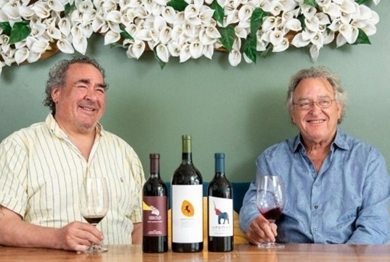 Two men smiling at a table with wine bottles and glasses, under white flowers against a green wall.