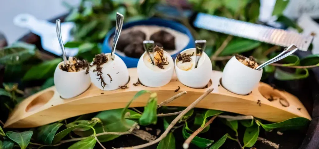 White appetizer cups with sprigs and sauces on a wooden board, surrounded by green leaves