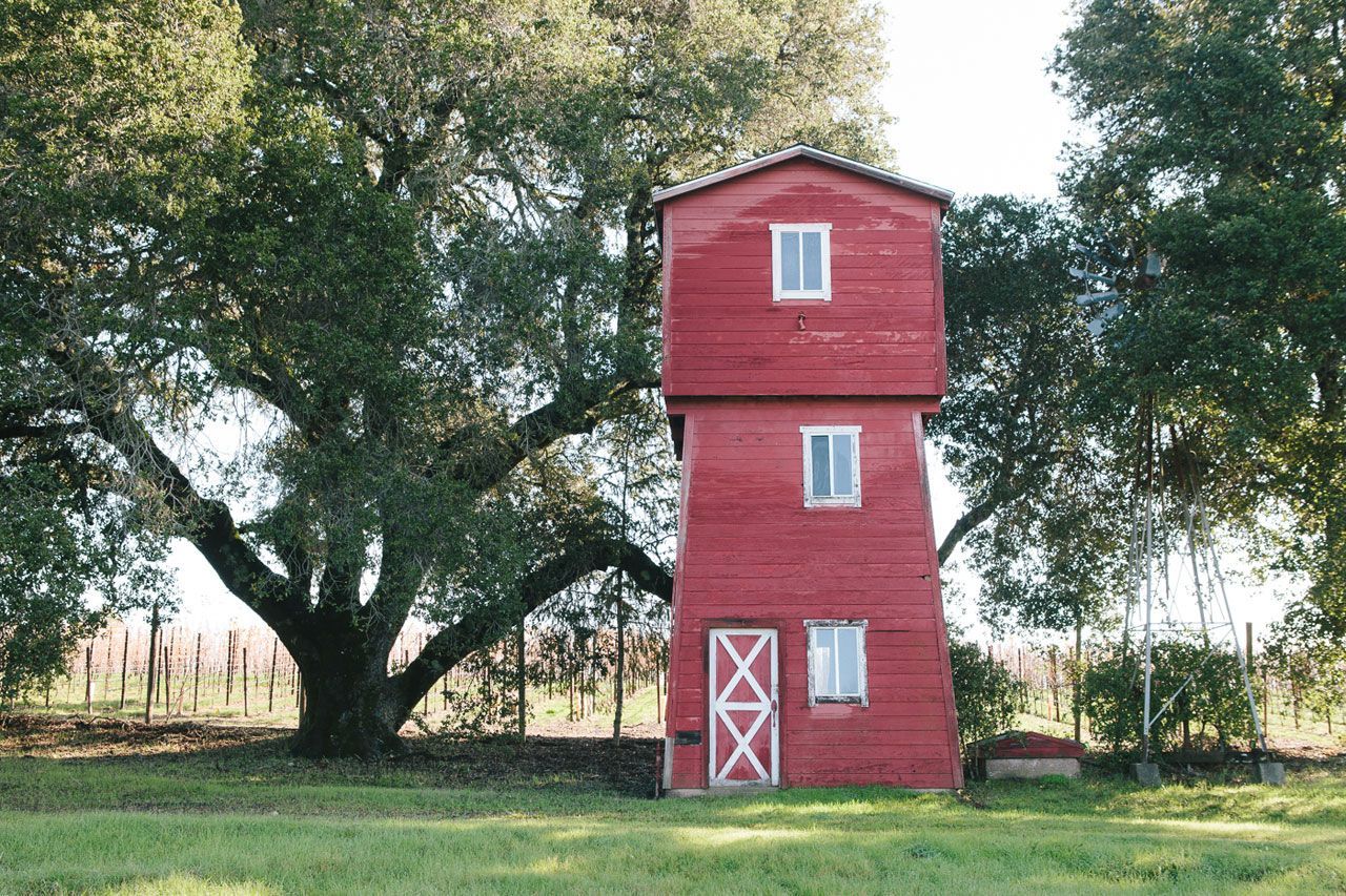 Red two-story tower-like shed with white windows and door in a grassy field under trees