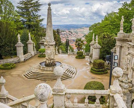 Stone terrace and ornate garden overlooking a city, with an obelisk fountain and statues amid trees and balustrades