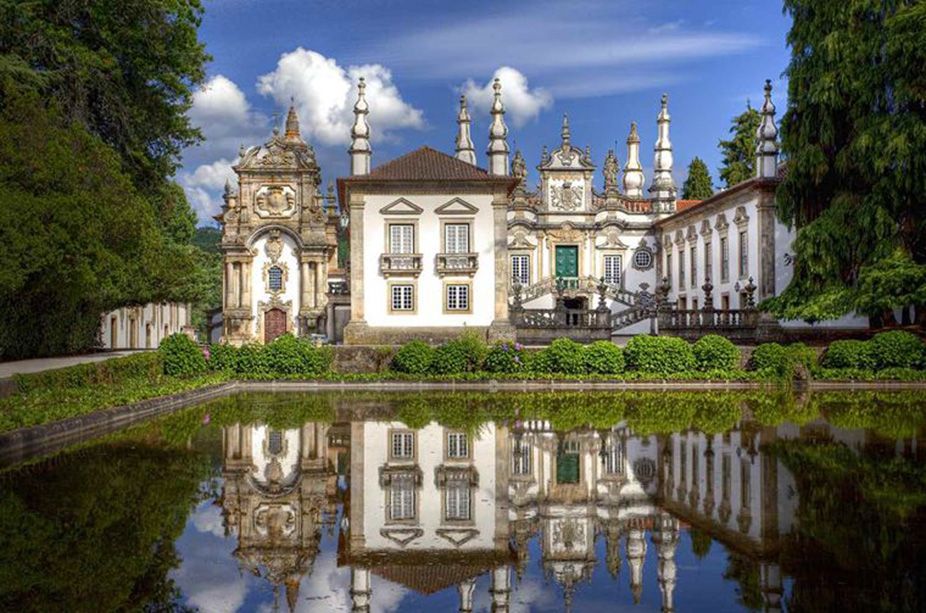 White palace with ornate towers reflected in a calm pond, framed by lush trees and blue sky.