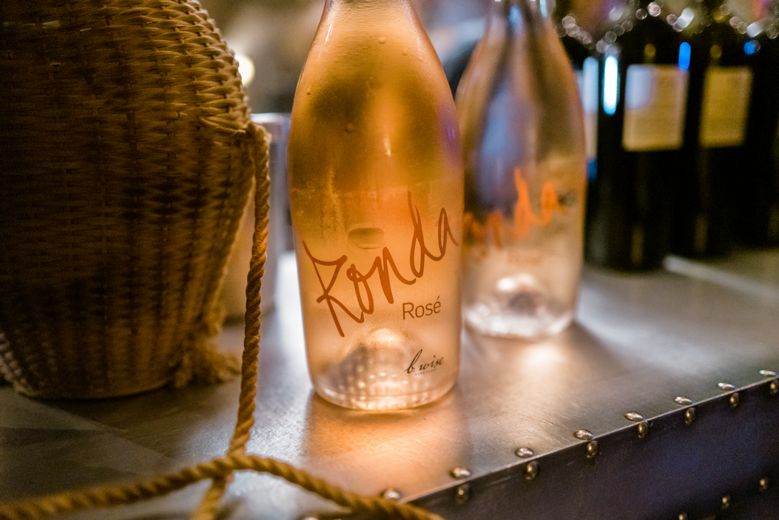 Raspberry rosé bottle on a metal table beside a woven chair, with other wine bottles blurred in the background