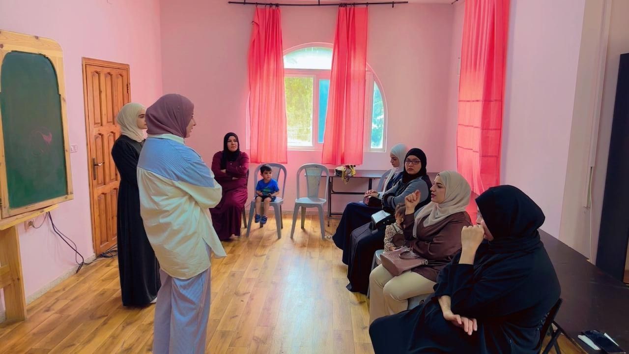 Small group meeting in a bright room, with seated attendees and a speaker near a chalkboard and pink curtains.