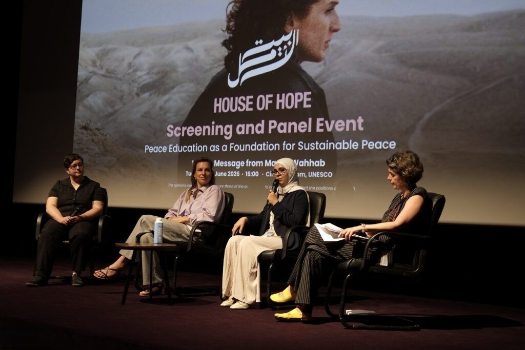Panel discussion on stage in front of “House of Hope” projection, with four speakers seated in chairs