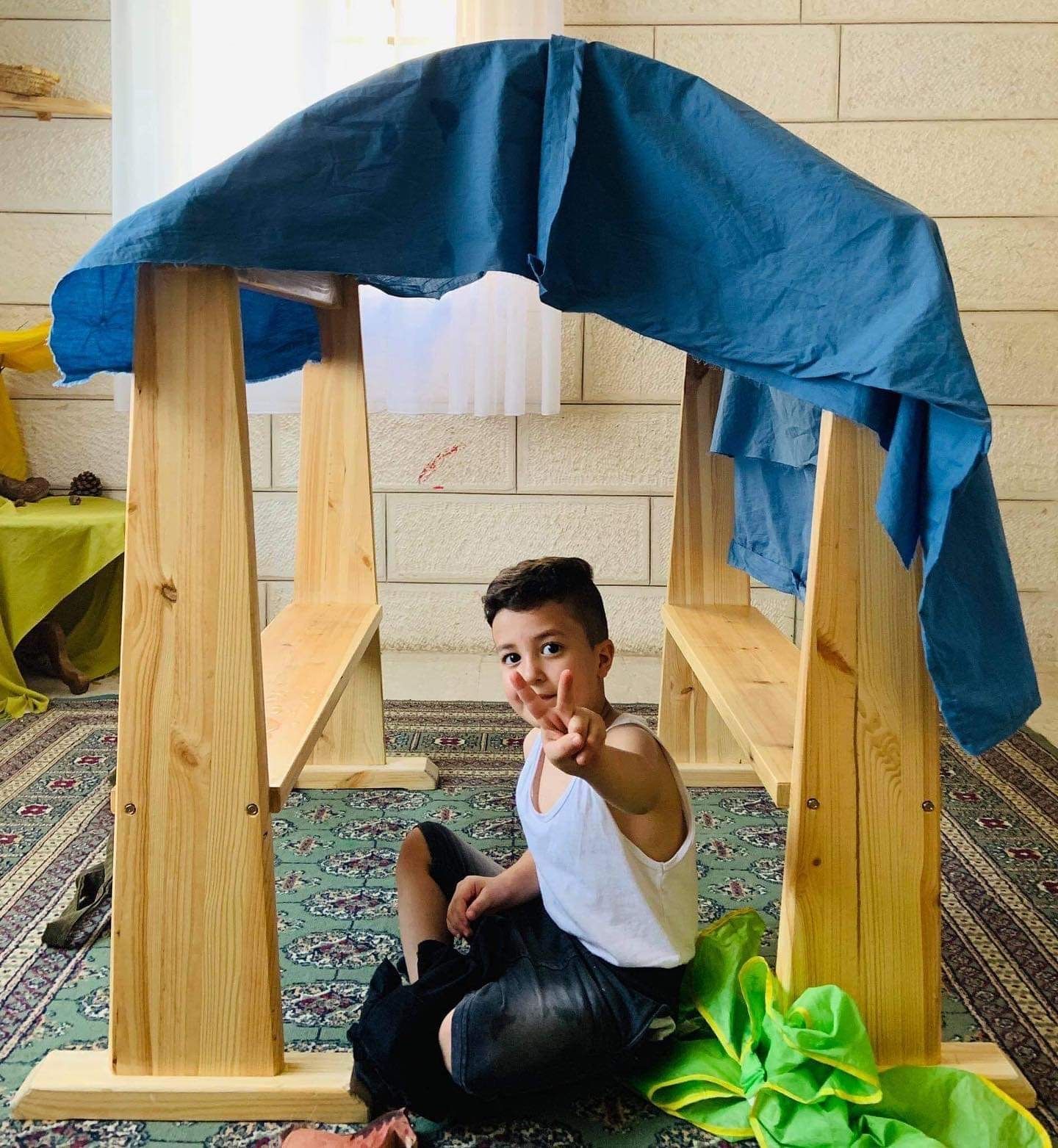 Child sitting inside a small wooden fort with a blue cloth roof, making a peace sign at the camera.