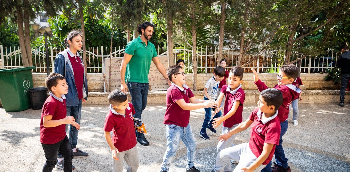 An adult in a green shirt supervises children playing a game in a tree-lined outdoor schoolyard.