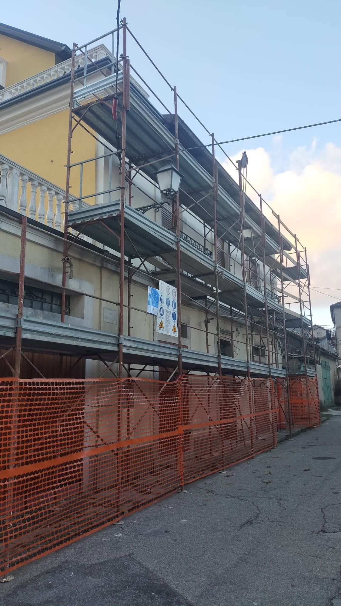 Impalcature attorno a un edificio giallo in una strada stretta, con reti di sicurezza arancioni lungo il marciapiede.