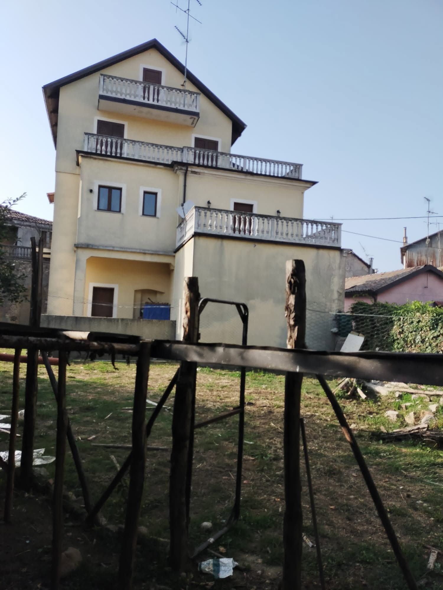 Casa beige a tre piani con balconi, situata dietro una recinzione metallica in un cortile erboso.