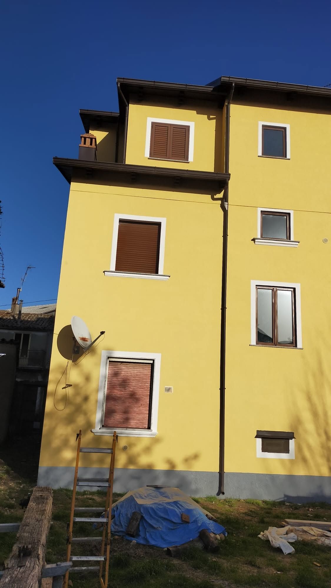Palazzo di appartamenti giallo con finestre con persiane marroni, parabola satellitare, scala e telone blu davanti.