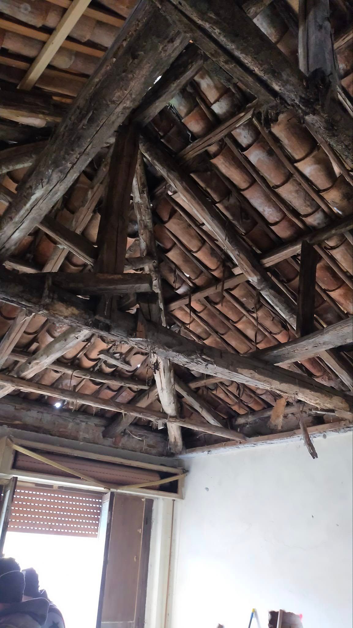Vista interna di una stanza rustica con travi in ​​legno a vista e tetto in tegole di terracotta, sopra una parete e una porta bianche.
