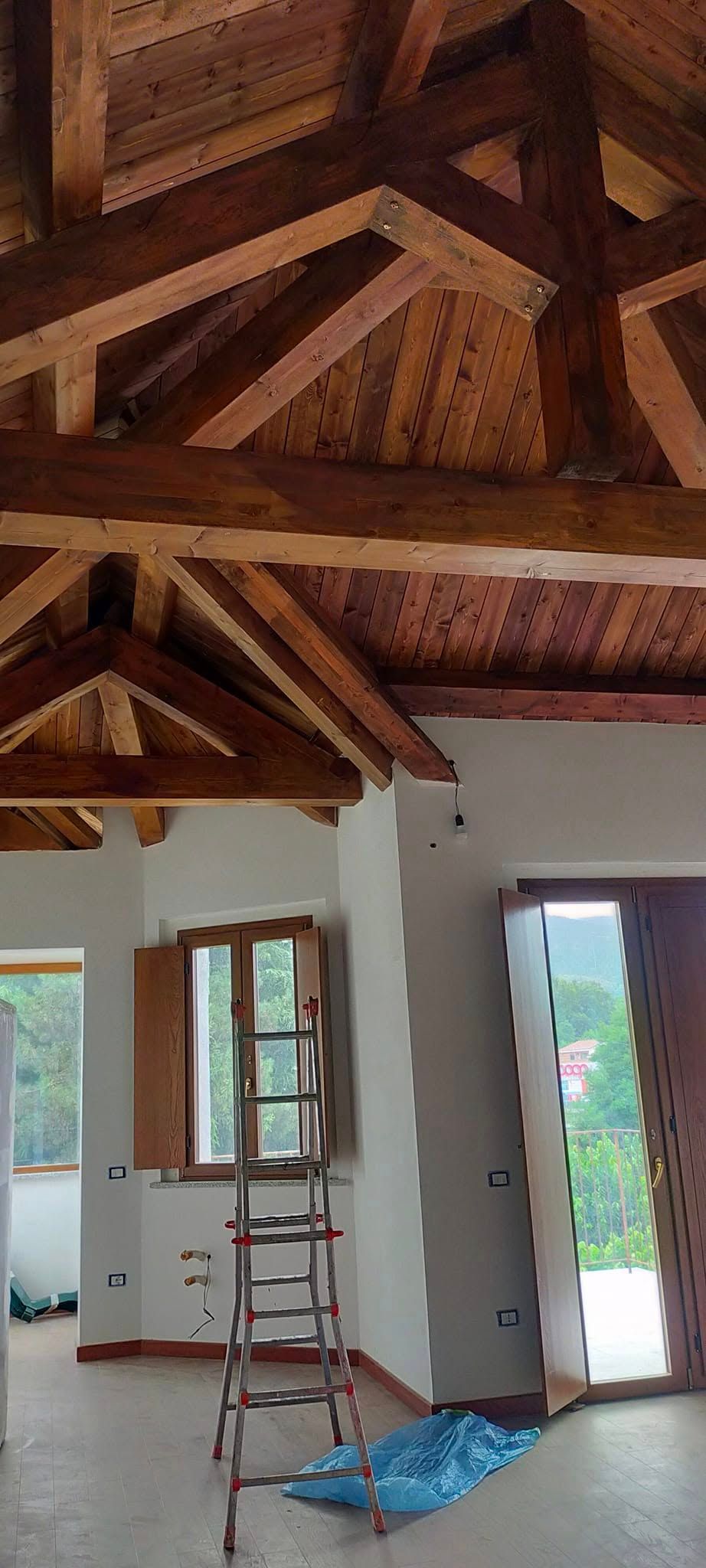 Interno con travi in ​​legno a vista sul soffitto, porta aperta, scala e materiali di ristrutturazione sul pavimento.