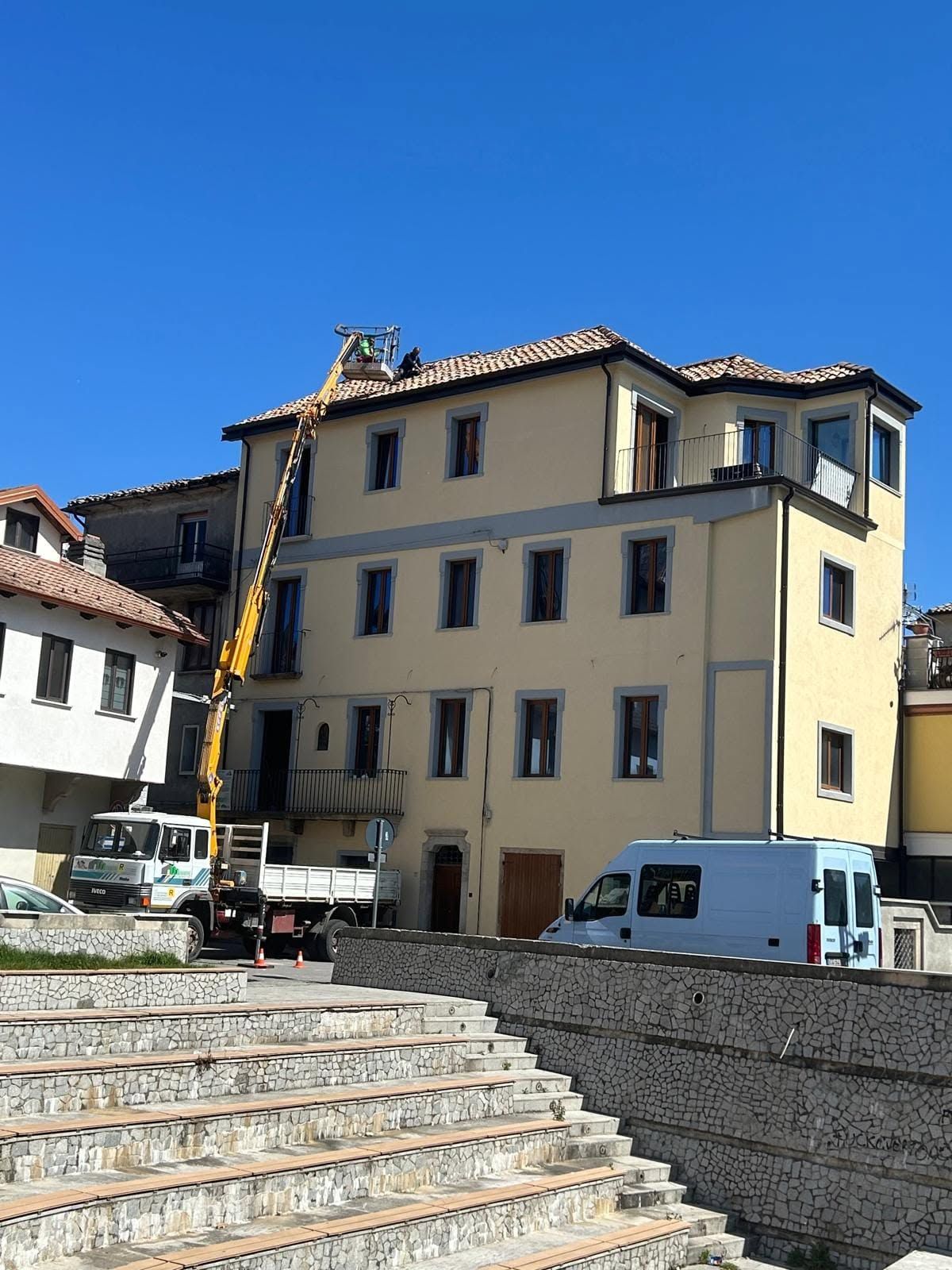 Una scalinata in pietra conduce a un edificio di colore giallo pallido, sotto un cielo azzurro limpido, dove sono parcheggiati dei furgoni.