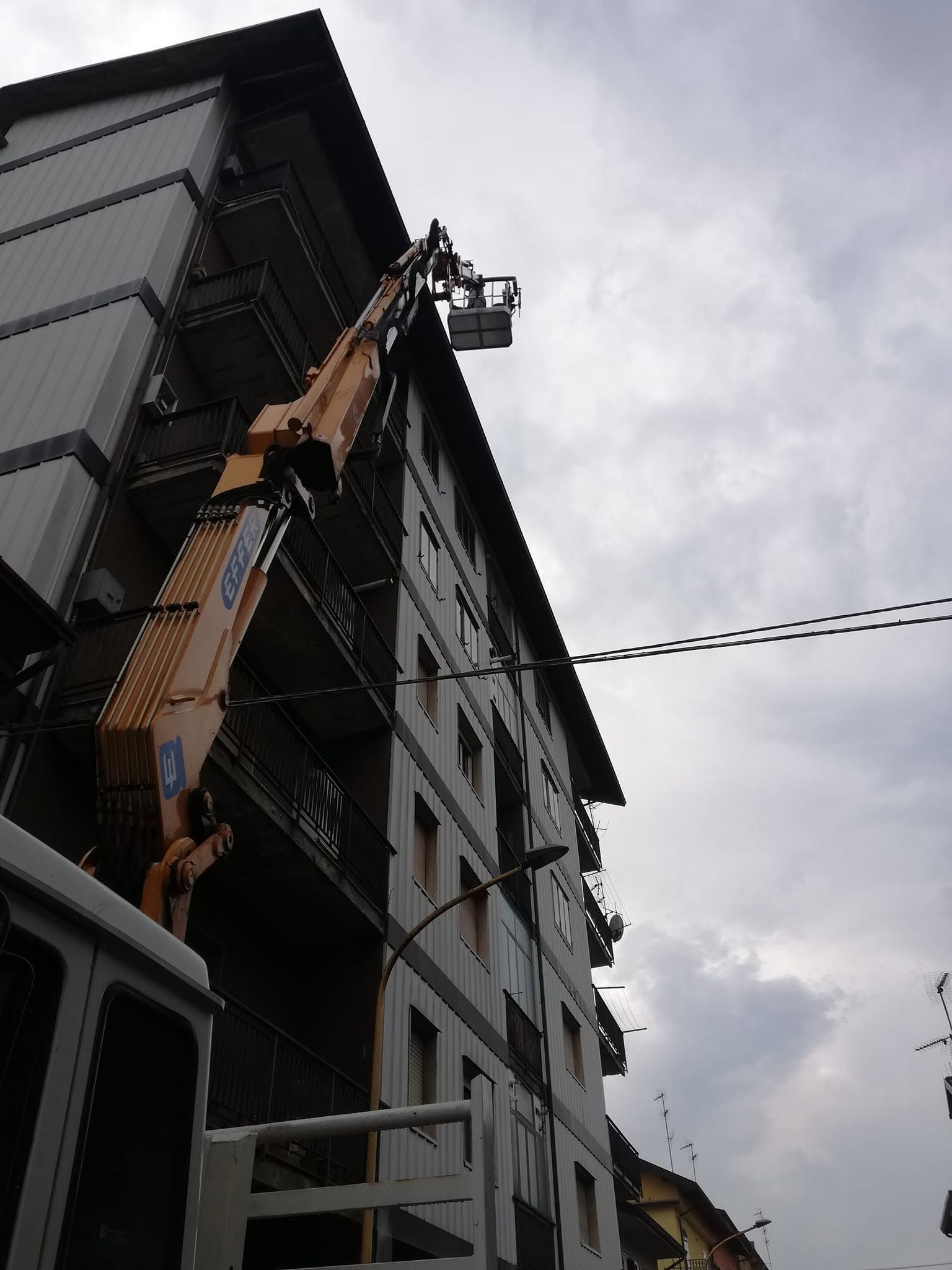 Una piattaforma elevatrice da cantiere si innalza lungo il fianco di un edificio grigio sotto un cielo nuvoloso.