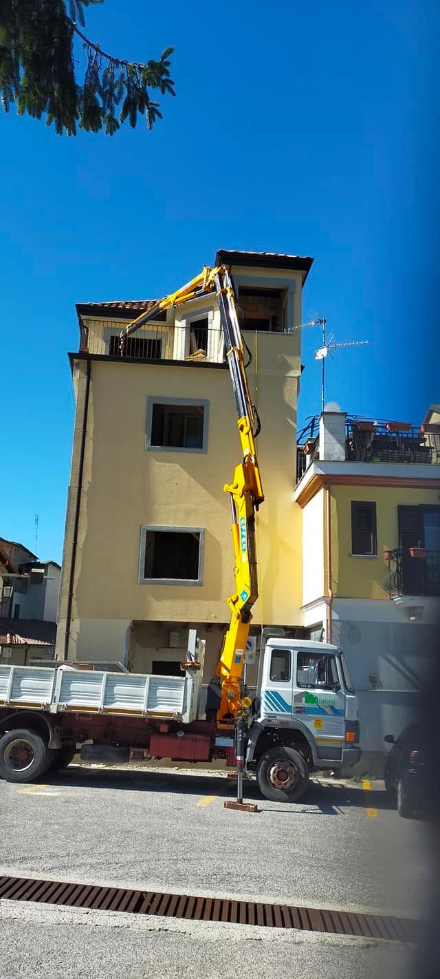 Un camion carico di detriti è parcheggiato accanto a un edificio giallo, sotto un cielo azzurro e limpido.