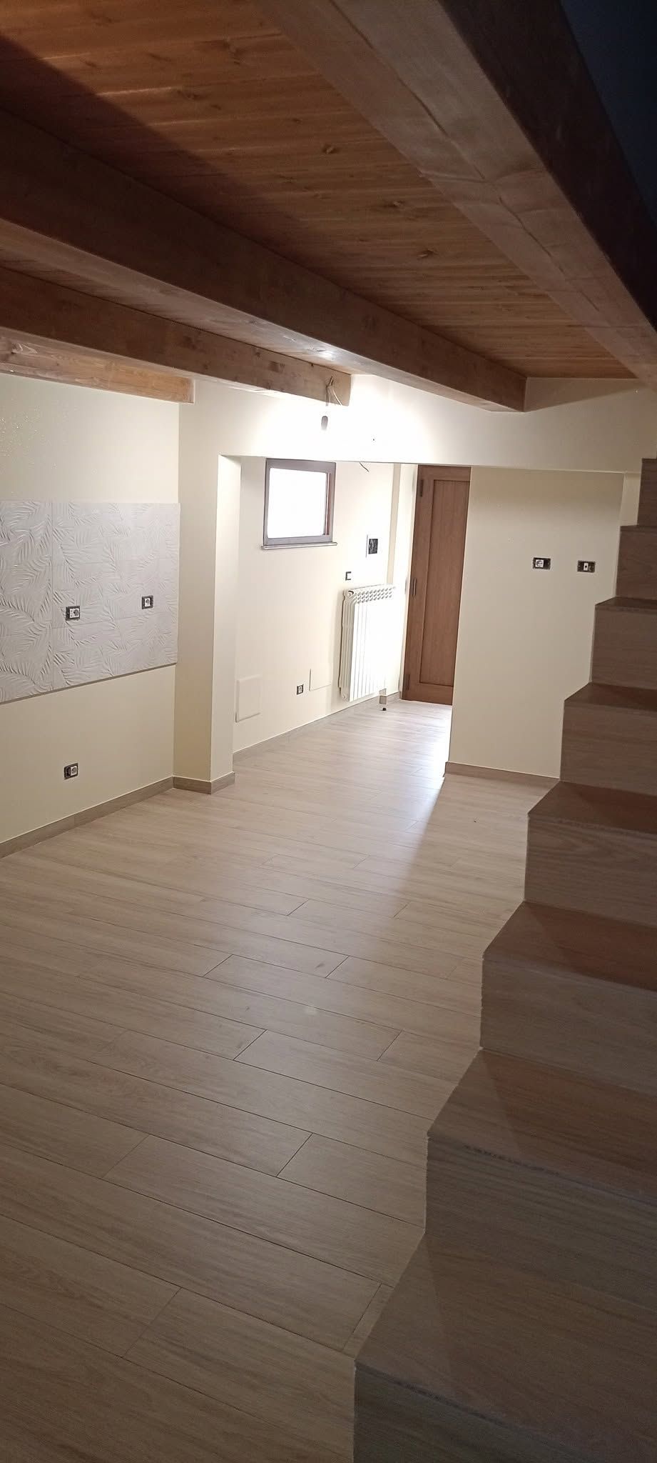 Corridoio vuoto e spoglio con travi a vista sul soffitto, scala e porta luminosa in fondo.