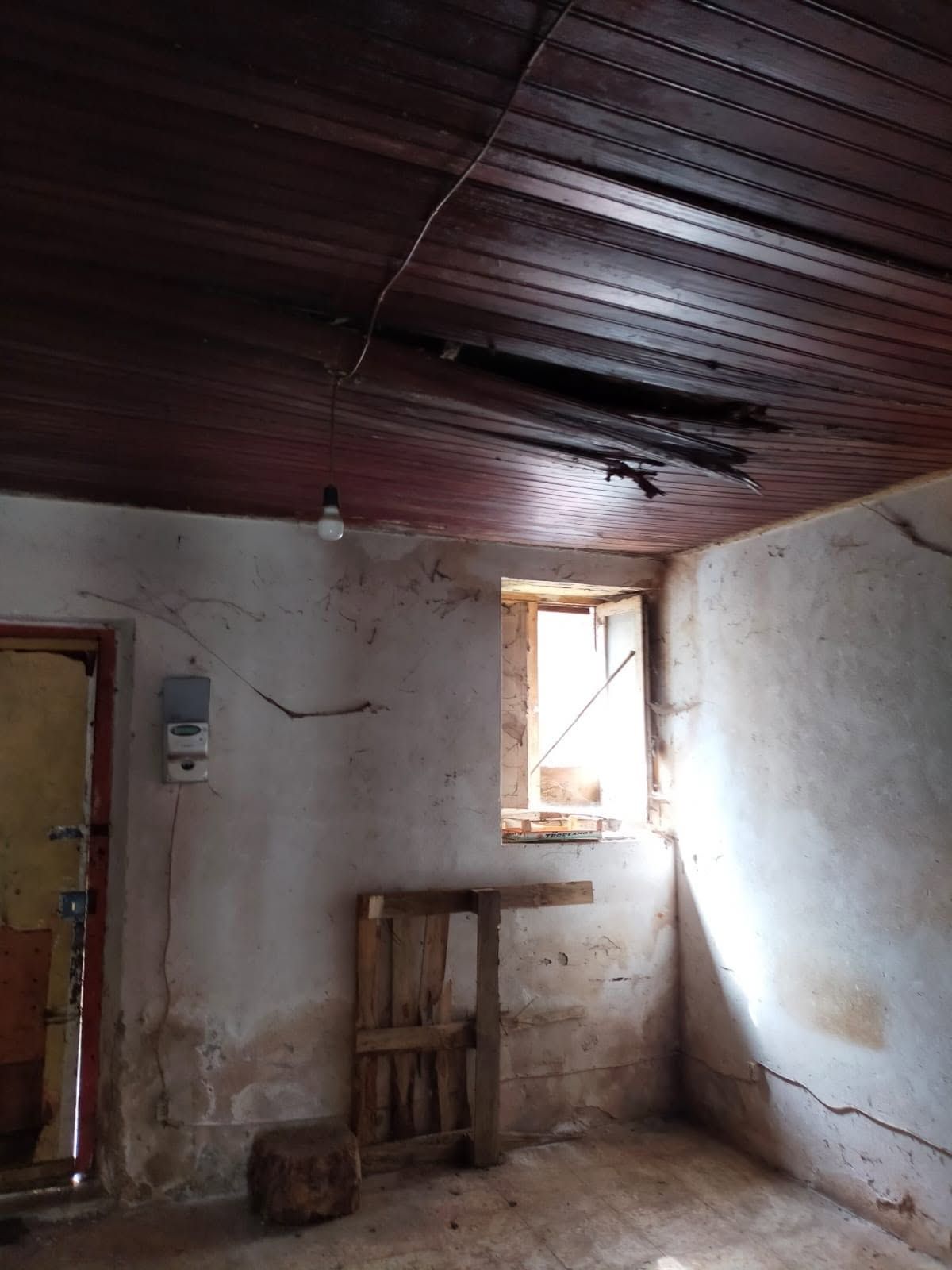 Piccola stanza spoglia con pareti bianche, soffitto in legno scuro, finestra aperta e una sedia di legno appoggiata alla parete.