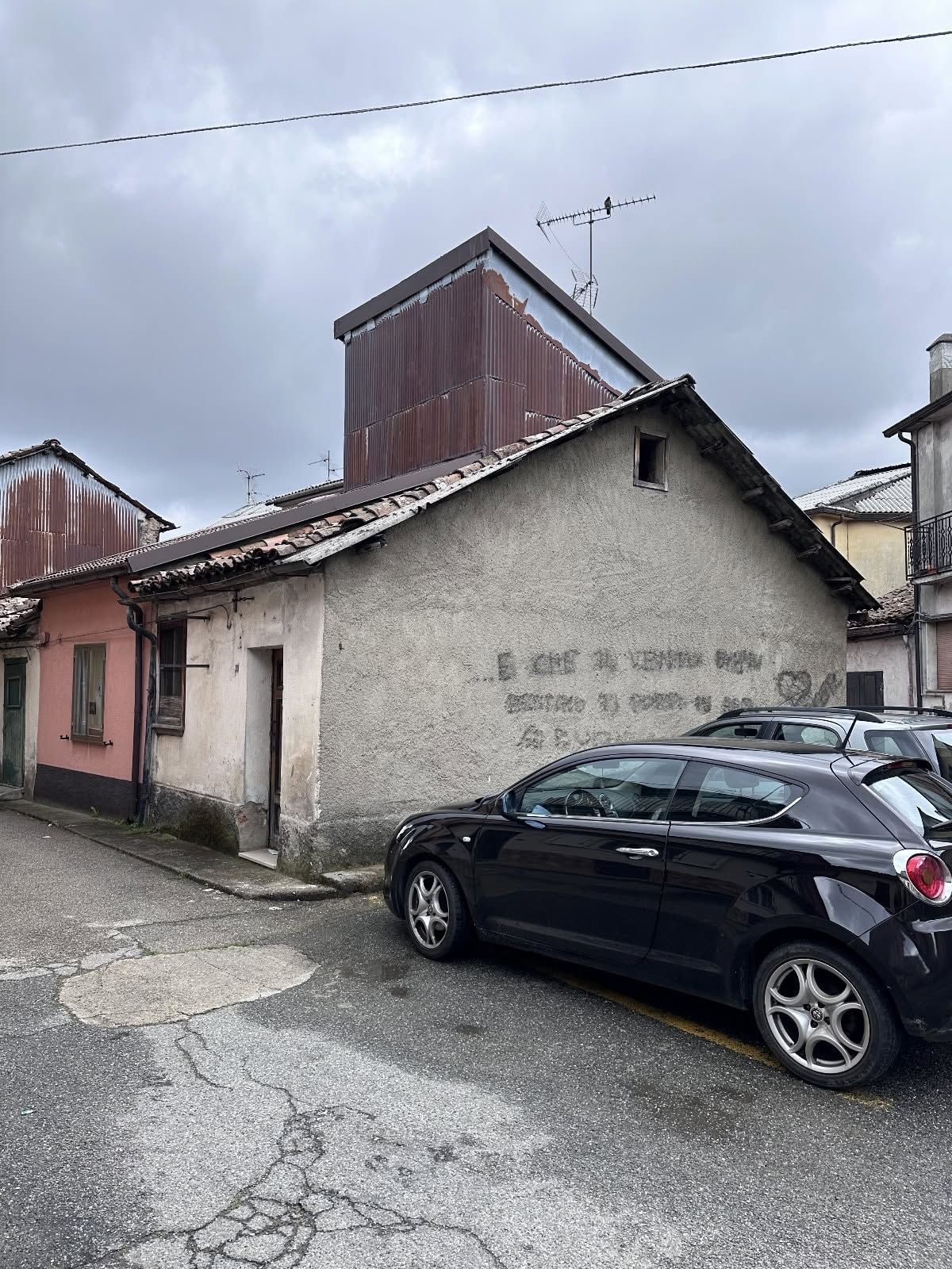Una strada stretta con vecchi edifici e un'auto nera parcheggiata accanto a un muro consumato dal tempo, sotto un cielo nuvoloso.