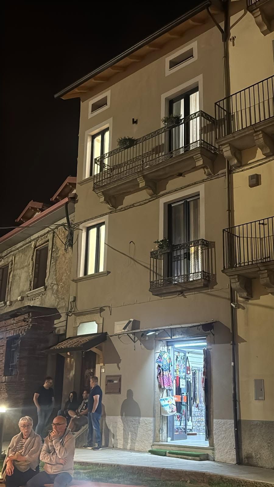 Scena notturna di strada con una vetrina illuminata sotto i balconi di un palazzo color beige, persone radunate all'esterno.