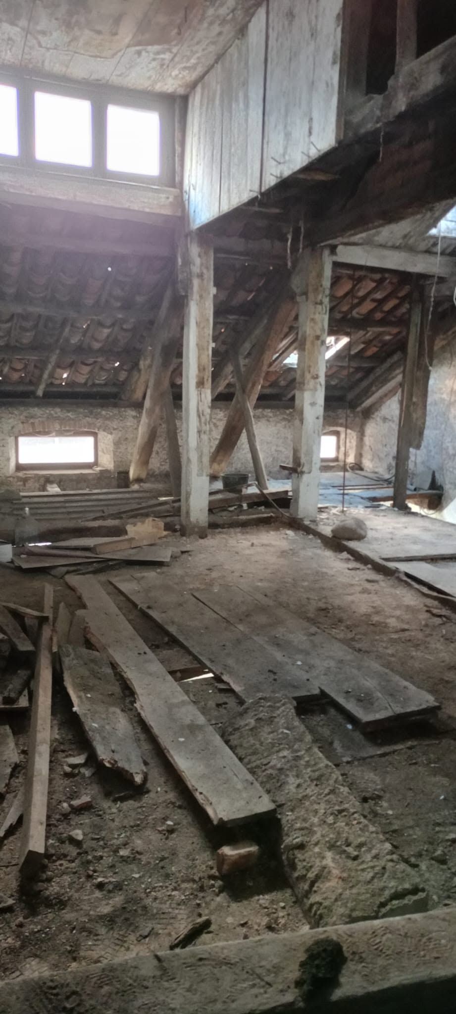Interno abbandonato con travi di legno rotte, detriti e finestre alte che lasciano entrare la luce del giorno.