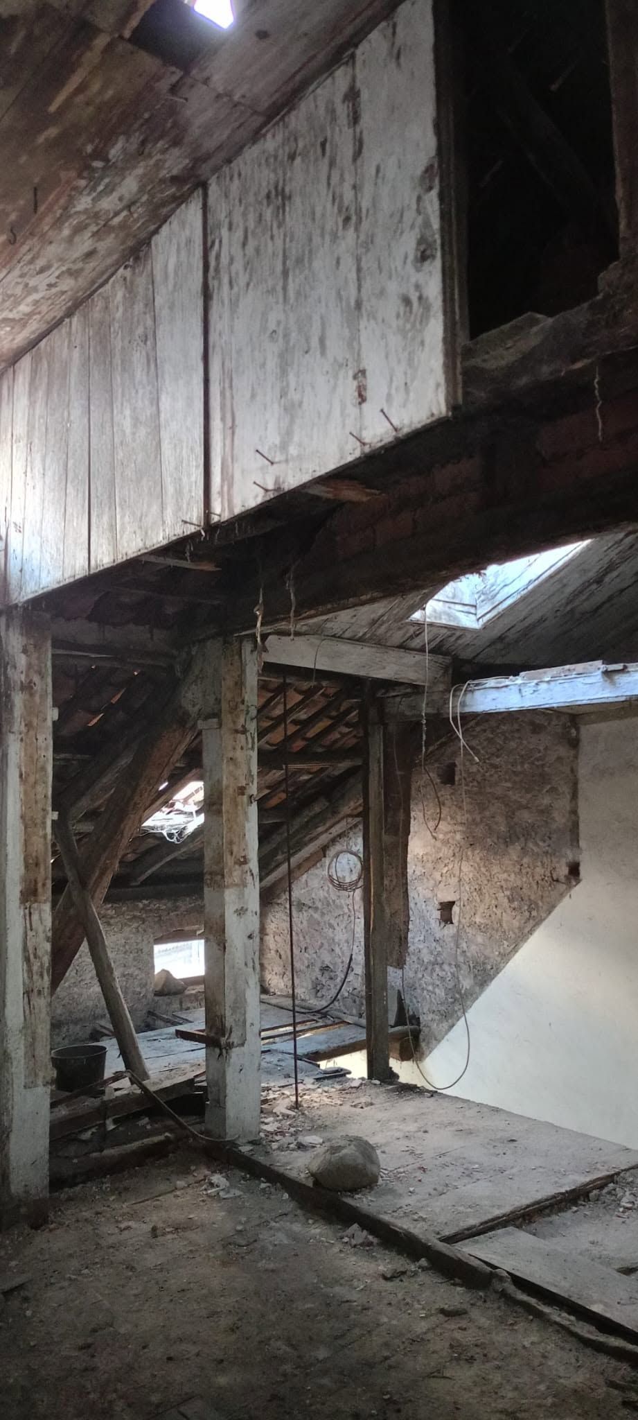 Interno abbandonato e danneggiato, con tetto rotto, travi di legno e detriti sul pavimento.