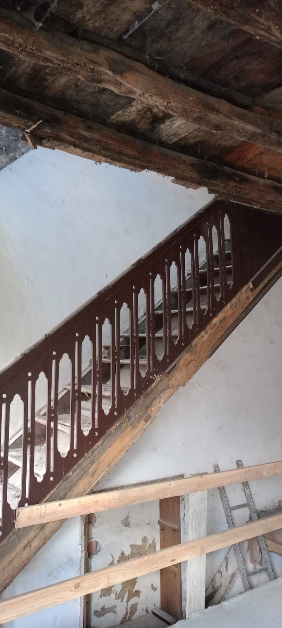 Scala interna in legno danneggiata sotto un soffitto inclinato con travi a vista.