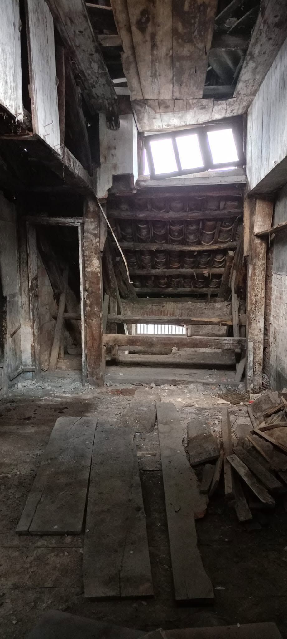 Interno abbandonato e in rovina, con scale rotte, detriti e luce del giorno che filtra attraverso una finestra danneggiata.