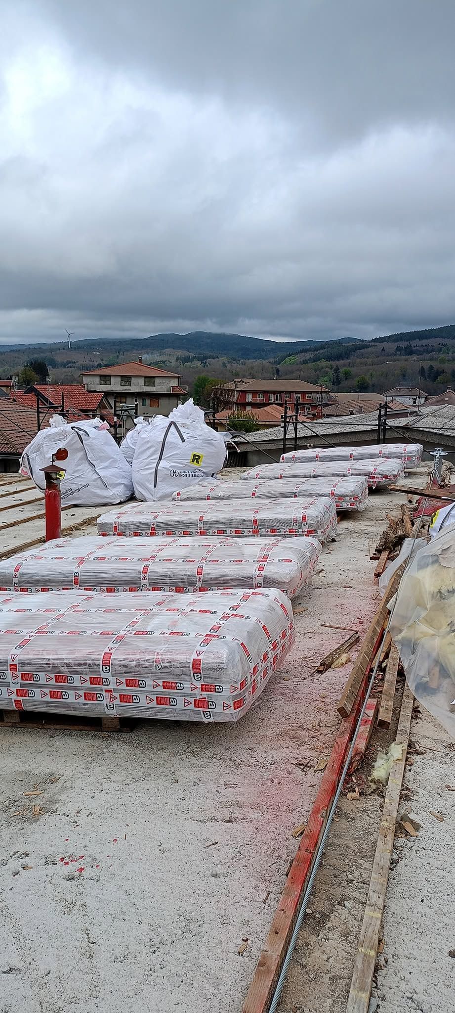 Tetto con pile di materiali imballati sotto un cielo nuvoloso, con vista su una città e sulle colline.