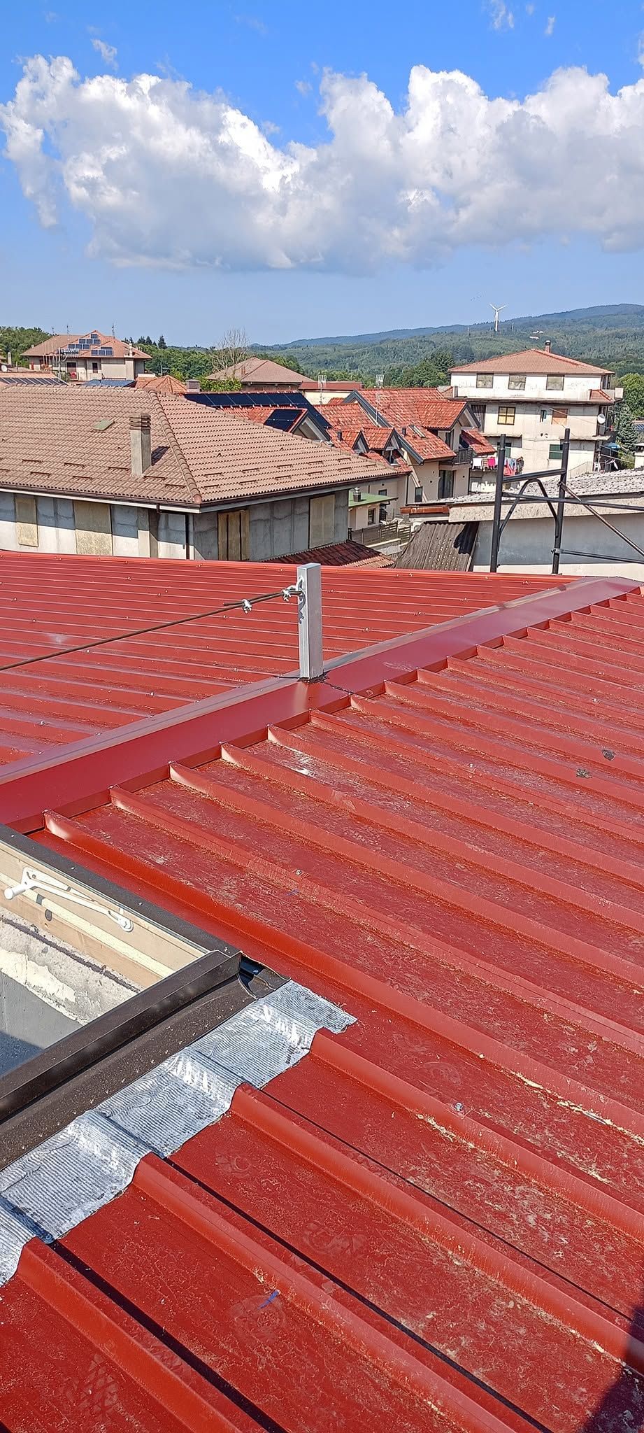 Tetti con lamiere rosse e tegole di terracotta che si affacciano su una città e sulle colline sotto un cielo nuvoloso.