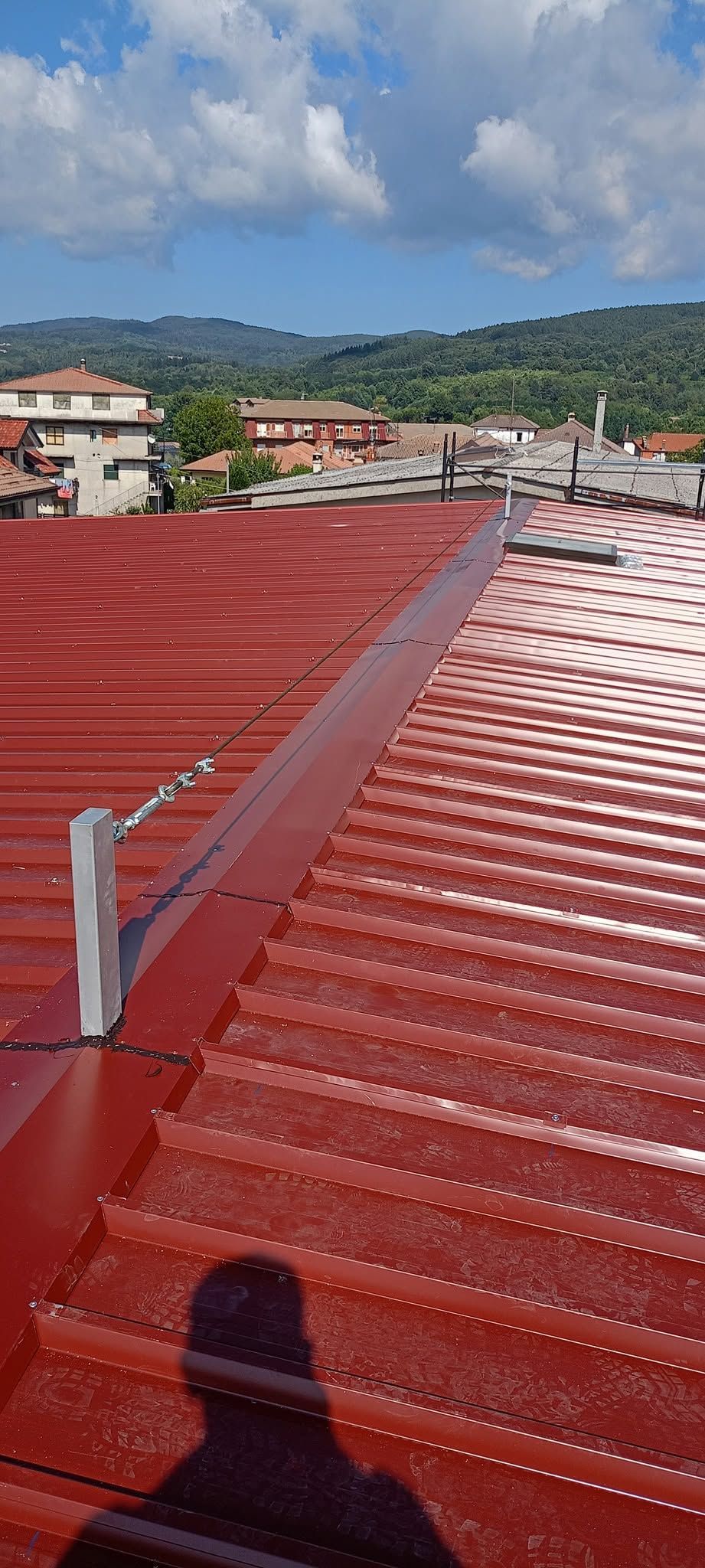 Tetto rosso ondulato con un'ombra in primo piano, che si affaccia su case e colline verdi sotto un cielo nuvoloso.