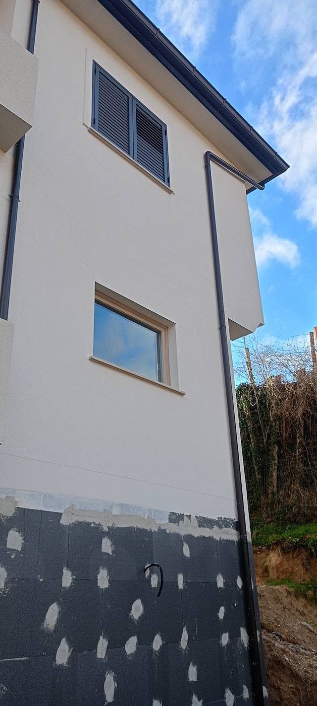 Vista esterna di una casa beige con finestre e muro inferiore scuro, sullo sfondo di un cielo azzurro.