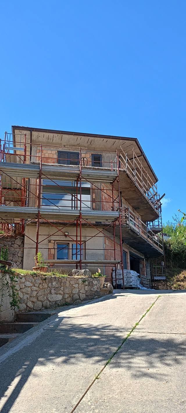 Casa in pietra in fase di ristrutturazione con impalcature accanto a un vialetto soleggiato e cielo azzurro.