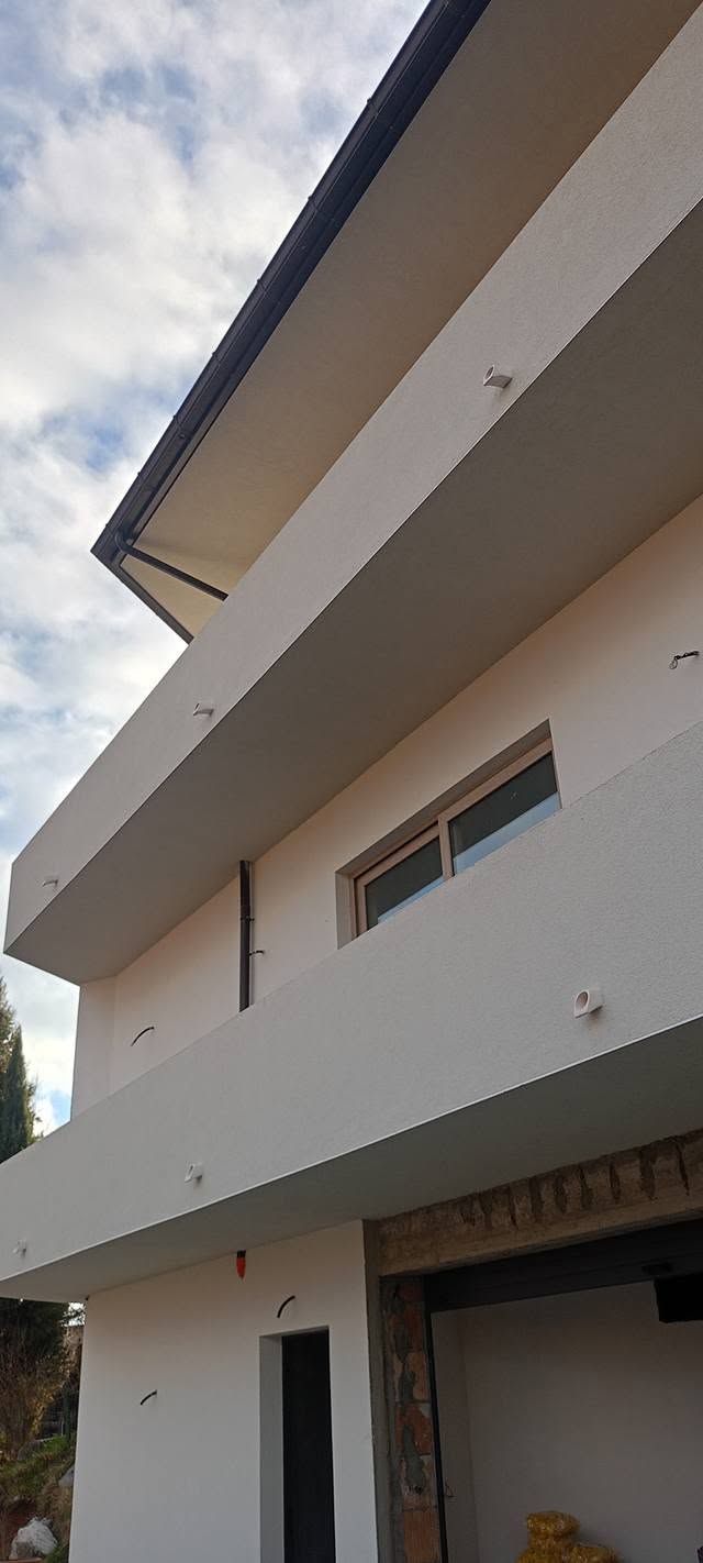 Esterno di una moderna casa bianca con tetto spiovente, balcone e finestre sotto un cielo nuvoloso.