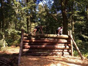 Two people are jumping over a log wall in the woods.