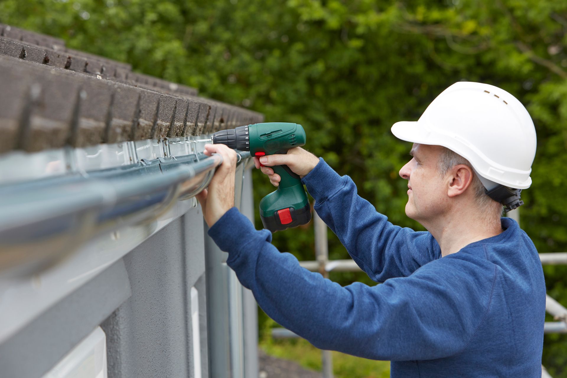 A man wearing a hard hat is working on a gutter with a drill.