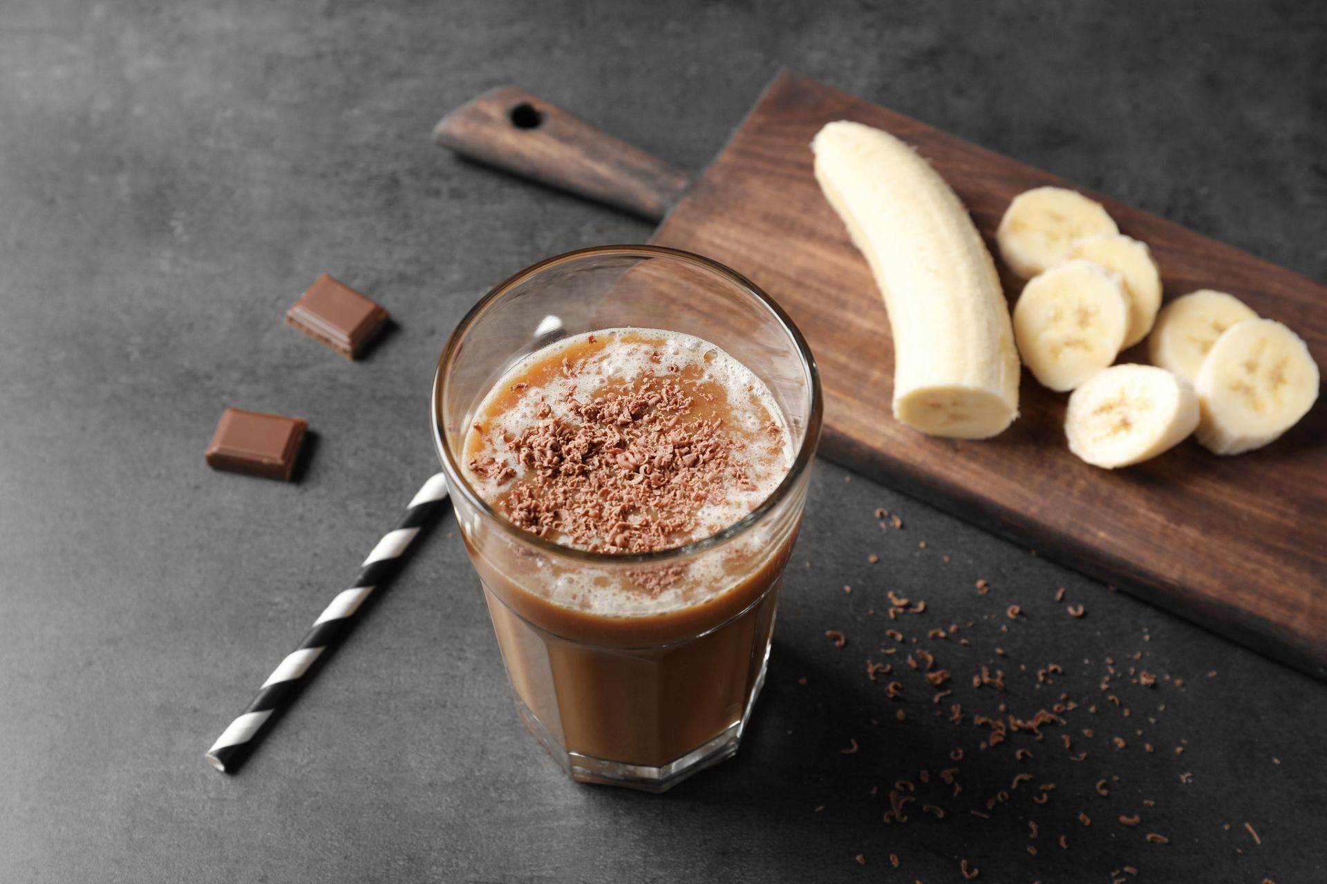 Chocolate banana smoothie in a glass, grated chocolate topping, with banana slices on a wooden board.