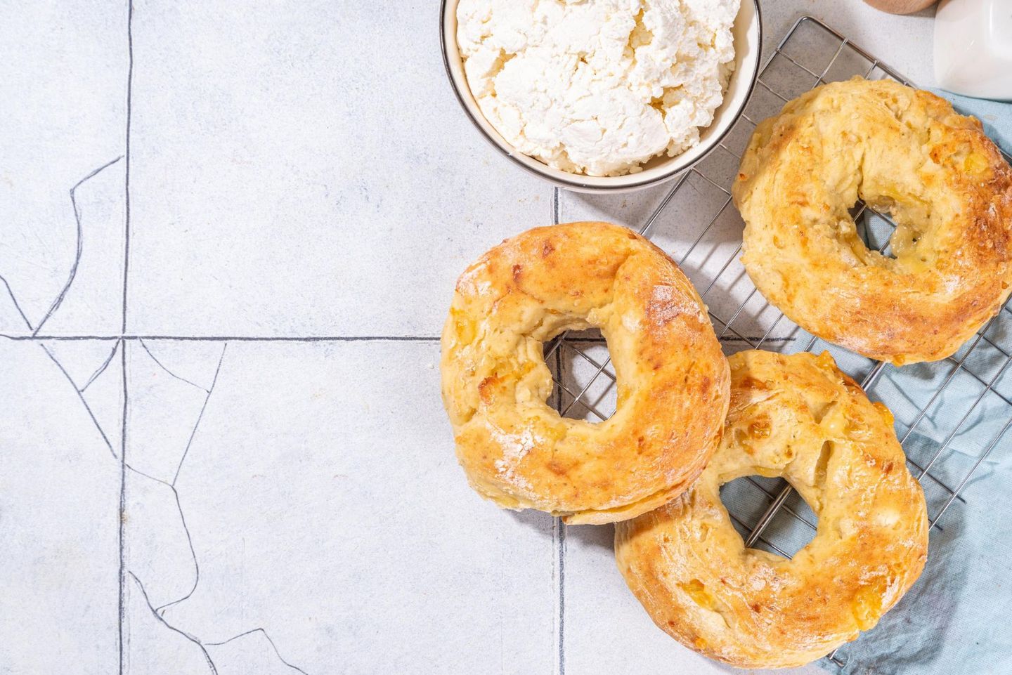 Three golden-brown bagels, a bowl of cream cheese, and a light blue cloth on a tiled surface.