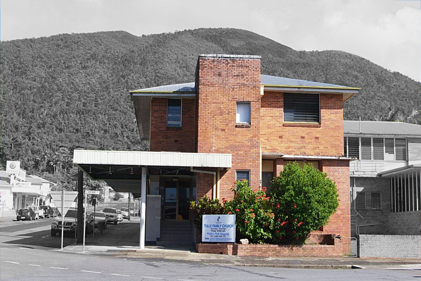 Small brick building with a covered entrance, set against hills and a street under a gray sky.