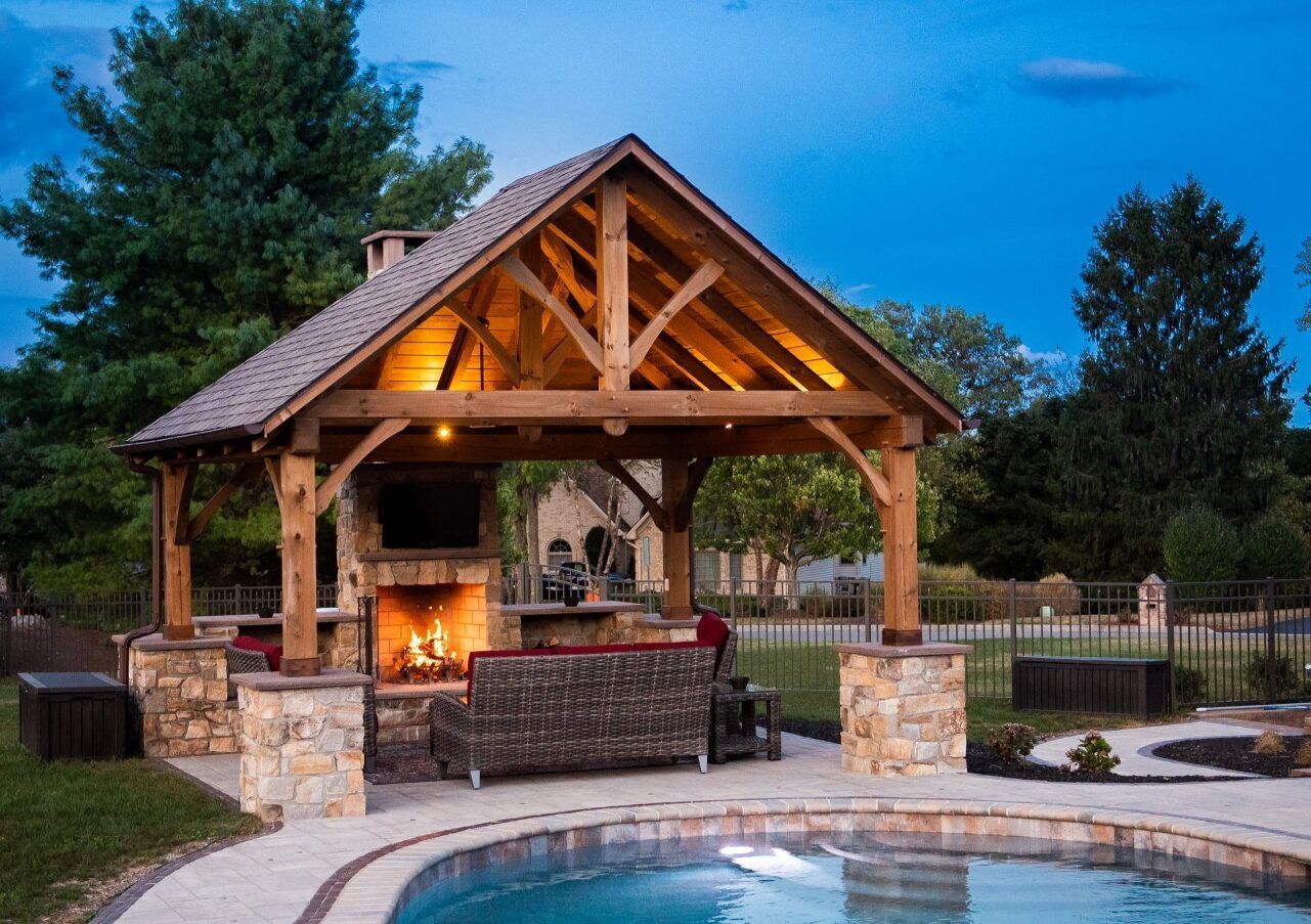 Covered outdoor patio with stone fireplace, wood beams, and seating beside a lit backyard pool at dusk