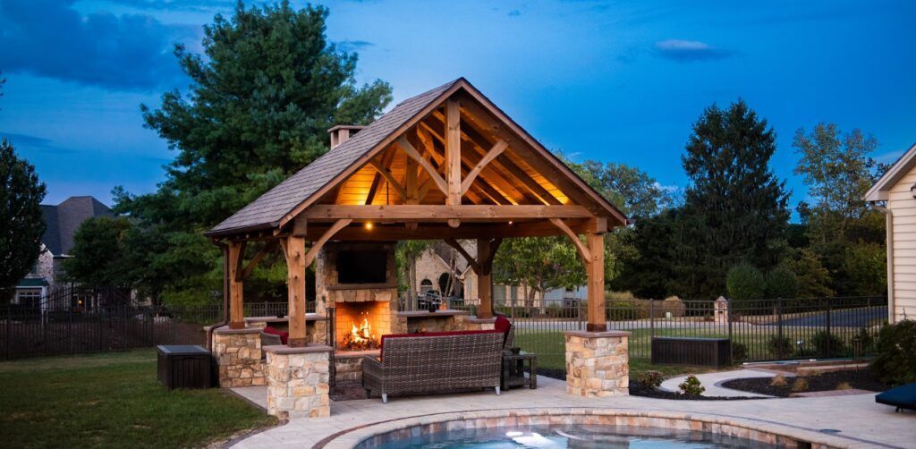 Stone pavilion with a peaked wooden roof and bench seating in a landscaped park at dusk