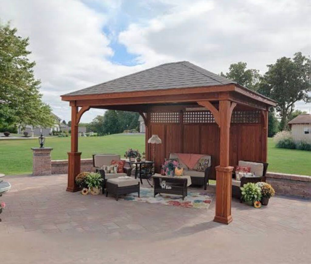 Wooden backyard gazebo with patio furniture, potted flowers, and a grassy park in the background