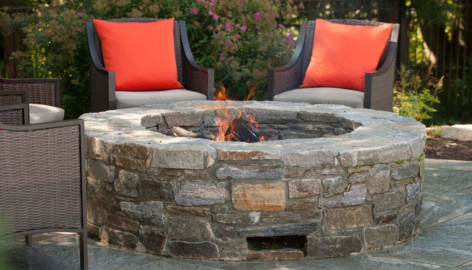 Stone fire pit with two gray chairs and bright orange cushions on a patio