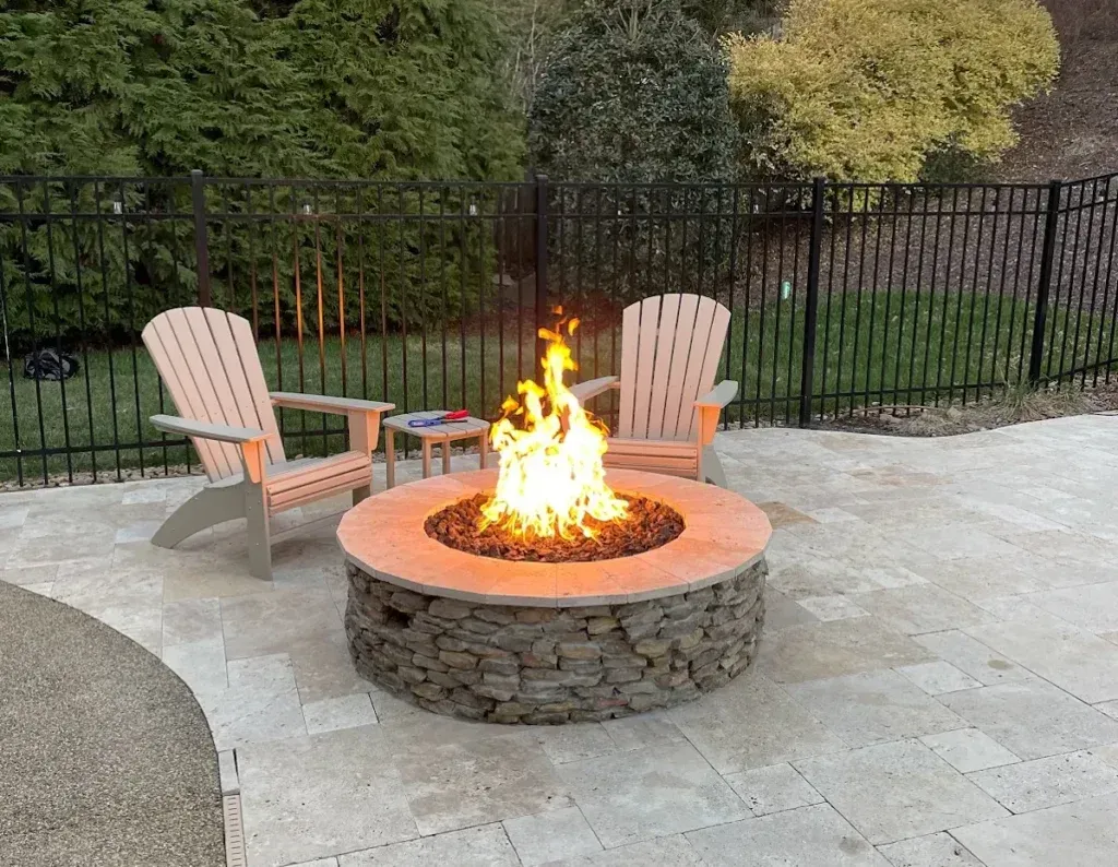 Backyard stone fire pit with flames, surrounded by two tan Adirondack chairs on a patio
