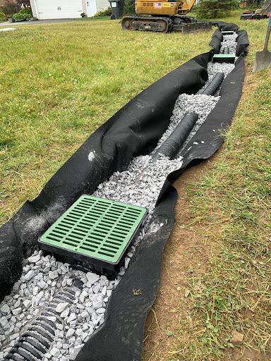 Landscape drainage trench with black fabric, gravel, and a green grating drain inlet